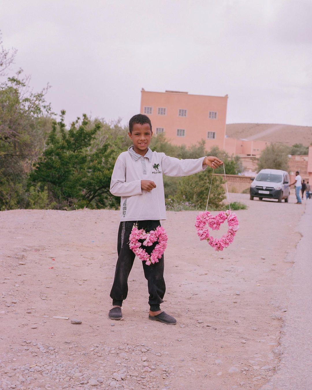 Boy selling heart-shaped floral decorations outdoors on a gravel road, with a building and vehicles in the background under an overcast sky.