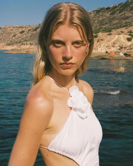 A blonde woman in a white halter top poses in front of a turquoise sea and rocky cliffs, creating a summery fashion portrait.