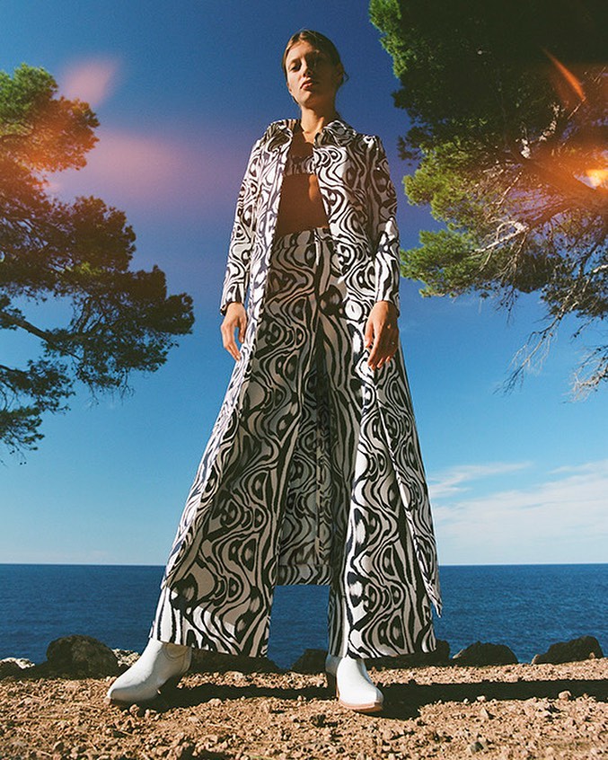Fashionable woman in a black and white patterned coat and pants stands outdoors near the ocean with a clear blue sky in the background.