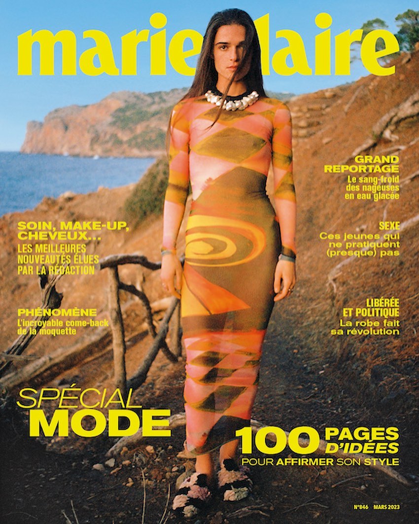 Marie Claire cover features a woman in an orange patterned dress standing in front of a rocky landscape near the sea on a sunny day.
