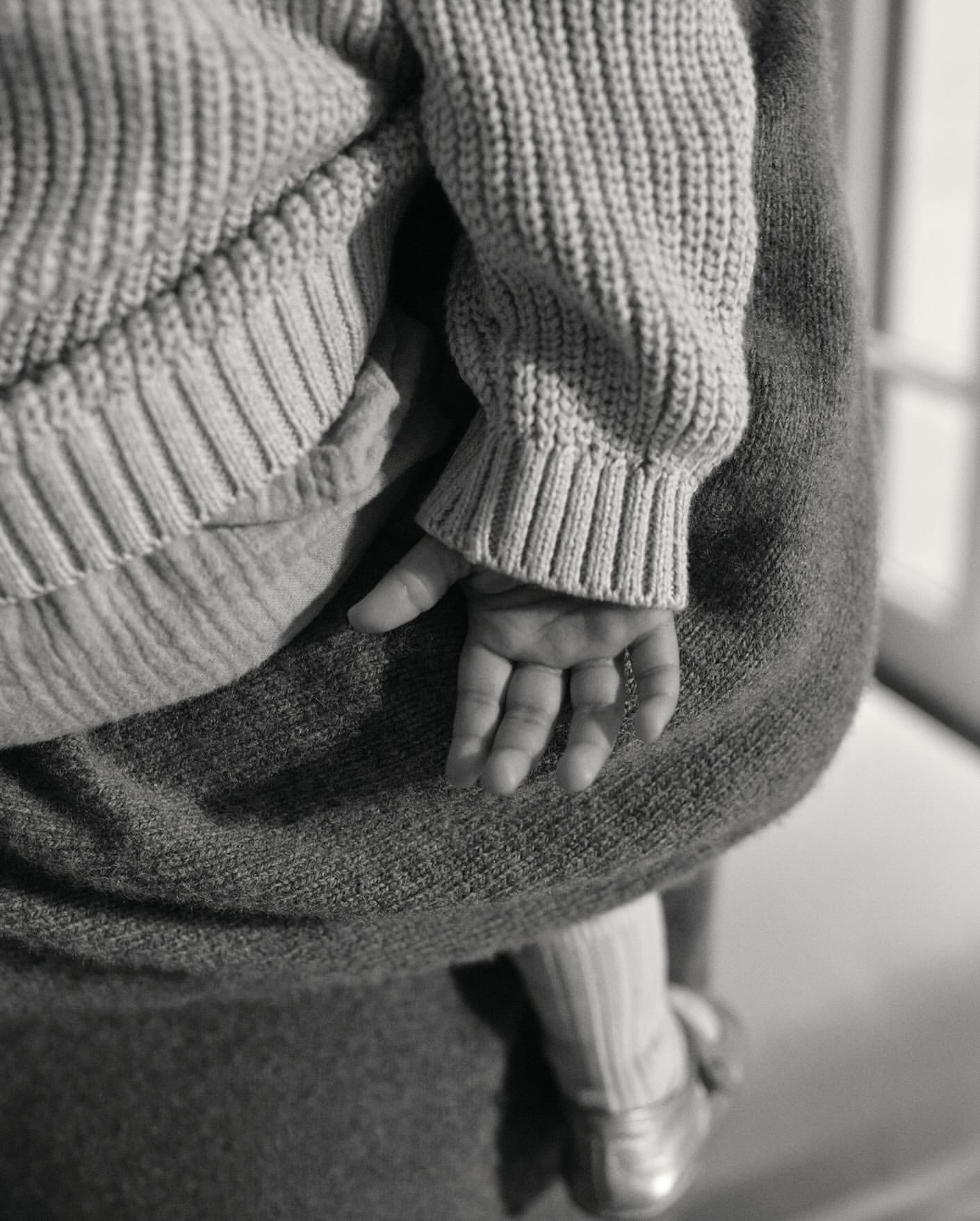 Close-up of a baby's hand and part of their body, being held by a person wearing gray clothing, emphasizing texture and gentle interaction.