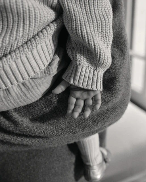 Close-up of a baby's hand and part of their body, being held by a person wearing gray clothing, emphasizing texture and gentle interaction.