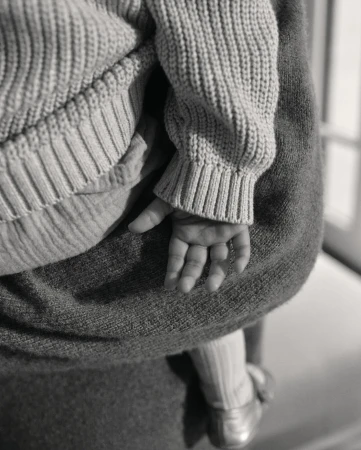 Close-up of a baby's hand and part of their body, being held by a person wearing gray clothing, emphasizing texture and gentle interaction.