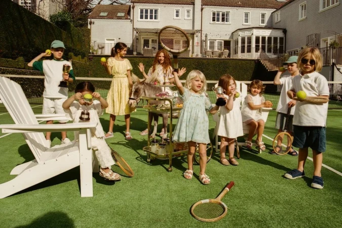 Group of children gather on a tennis court for an outdoor party with tea, tennis balls, rackets, and a small pony pulling a cart.