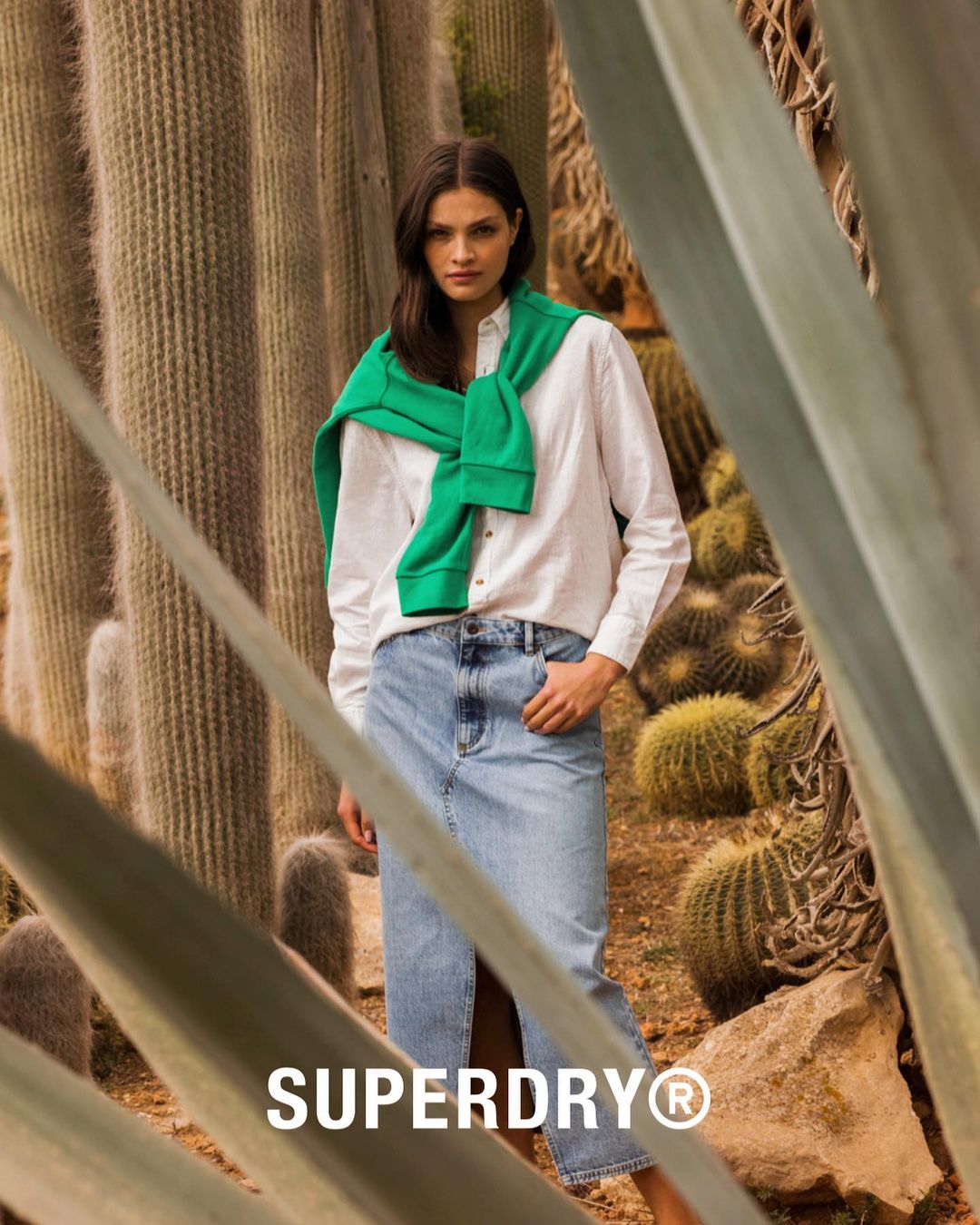 A woman wearing a white shirt and blue denim skirt stands among cacti, with a green sweater around her shoulders. The Superdry logo is visible.