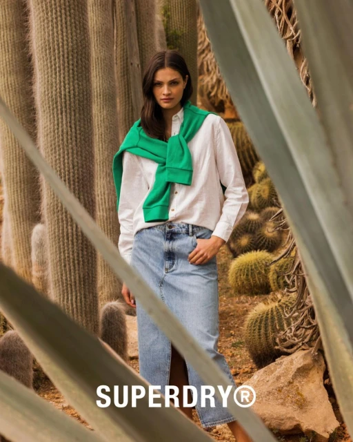 A woman wearing a white shirt and blue denim skirt stands among cacti, with a green sweater around her shoulders. The Superdry logo is visible.