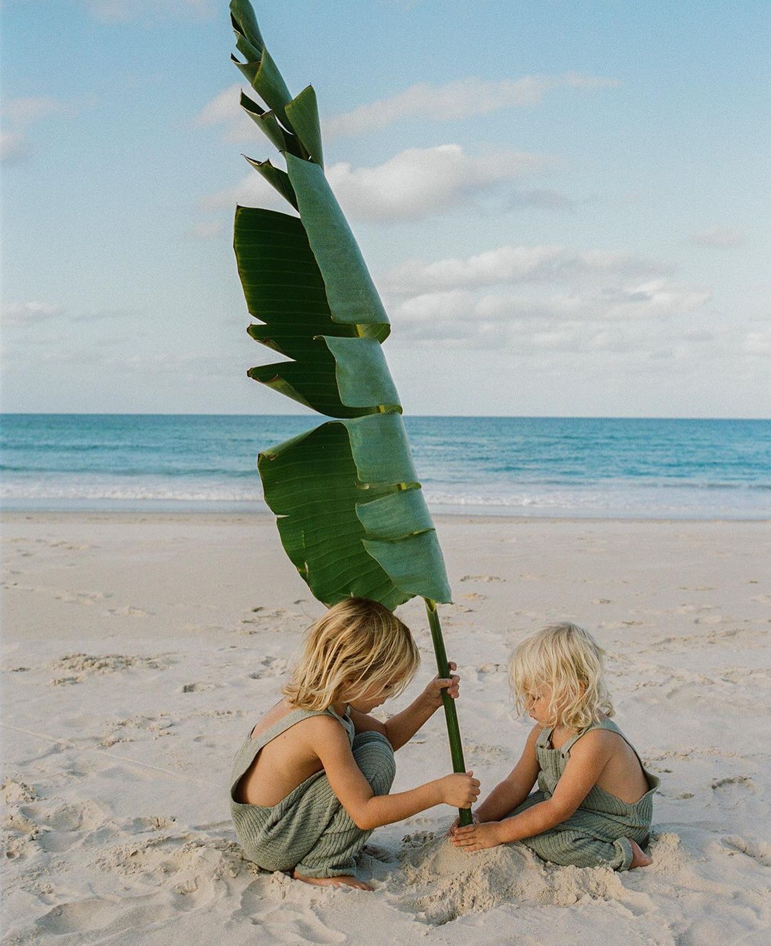 Two children play on a sandy beach with a large green leaf. The horizon shows the ocean under a blue sky with scattered clouds.