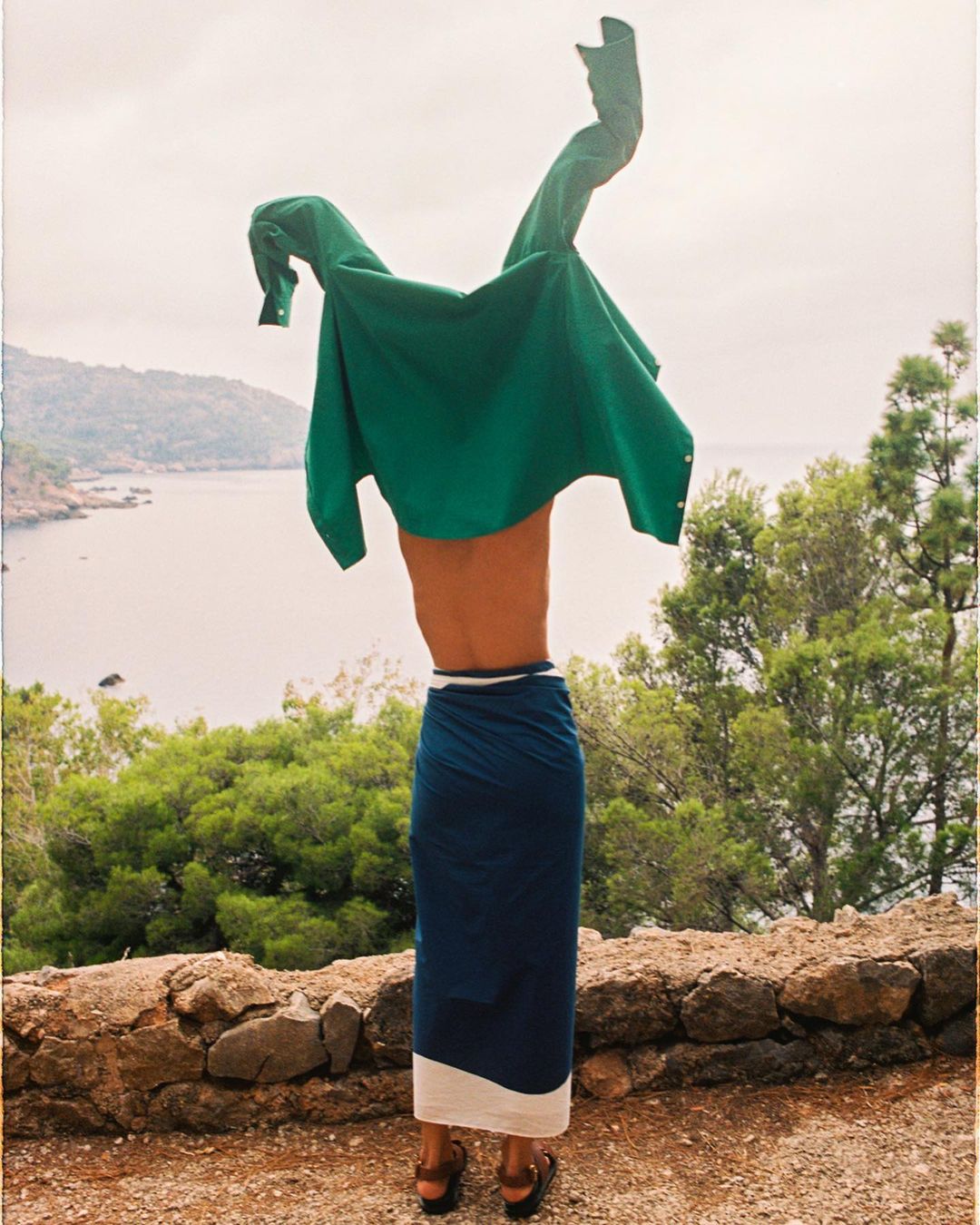 Person wearing a sarong on a stone ledge holding a green shirt in the air near trees with a distant view of the water.