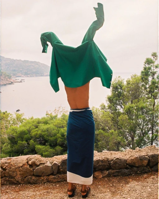 Person wearing a sarong on a stone ledge holding a green shirt in the air near trees with a distant view of the water.