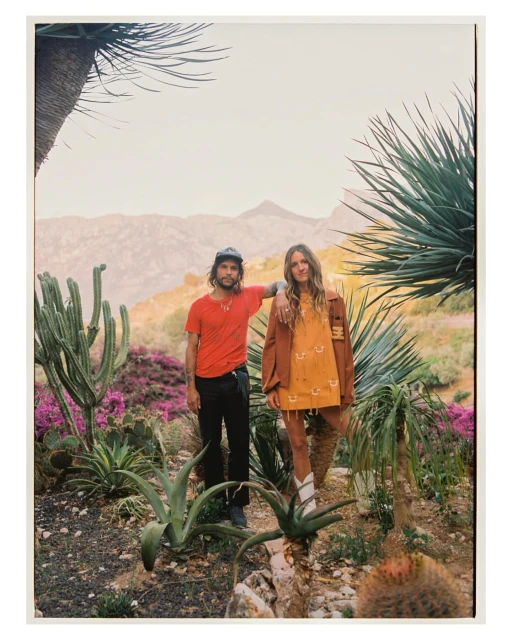Couple stands in a desert landscape with mountains in the background, surrounded by cacti, agave plants, and flowering purple bushes.