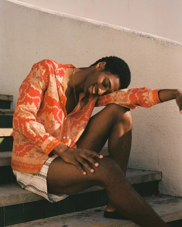 Man wearing a red patterned shirt and light shorts seated outdoors on concrete steps, smiling with his head resting on his arm.