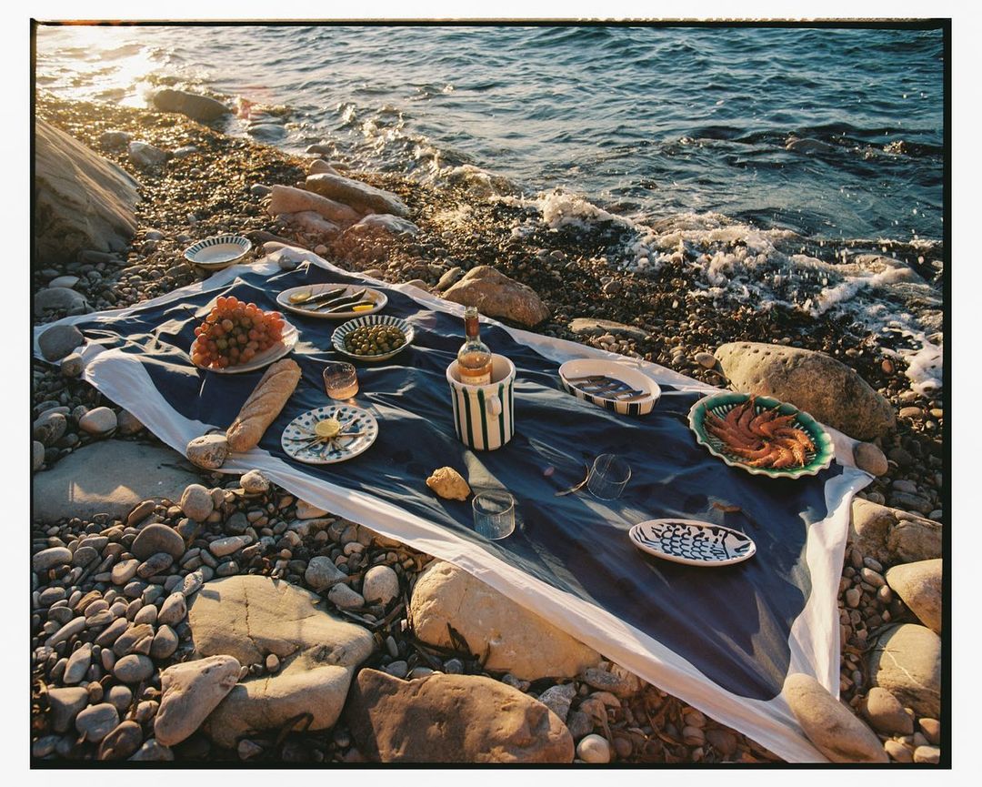 Outdoor picnic on a rocky beach with a blue and white tablecloth, featuring grapes, bread, and various dishes set near gentle ocean waves.