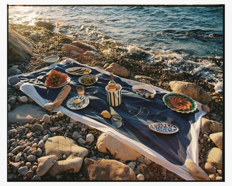 Outdoor picnic on a rocky beach with a blue and white tablecloth, featuring grapes, bread, and various dishes set near gentle ocean waves.