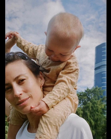 Mother carries her baby on her shoulders outside in a park setting, with a tall building visible in the background on a sunny day.