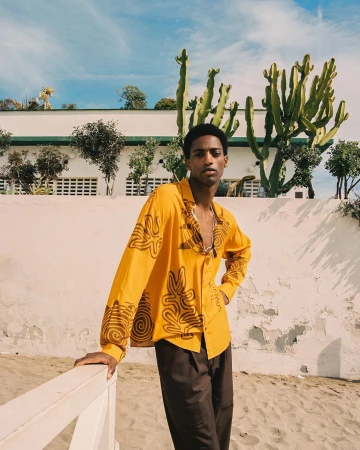 A young Black man wearing a yellow patterned button-up shirt leans against a white railing outdoors in front of tall cacti and a building.