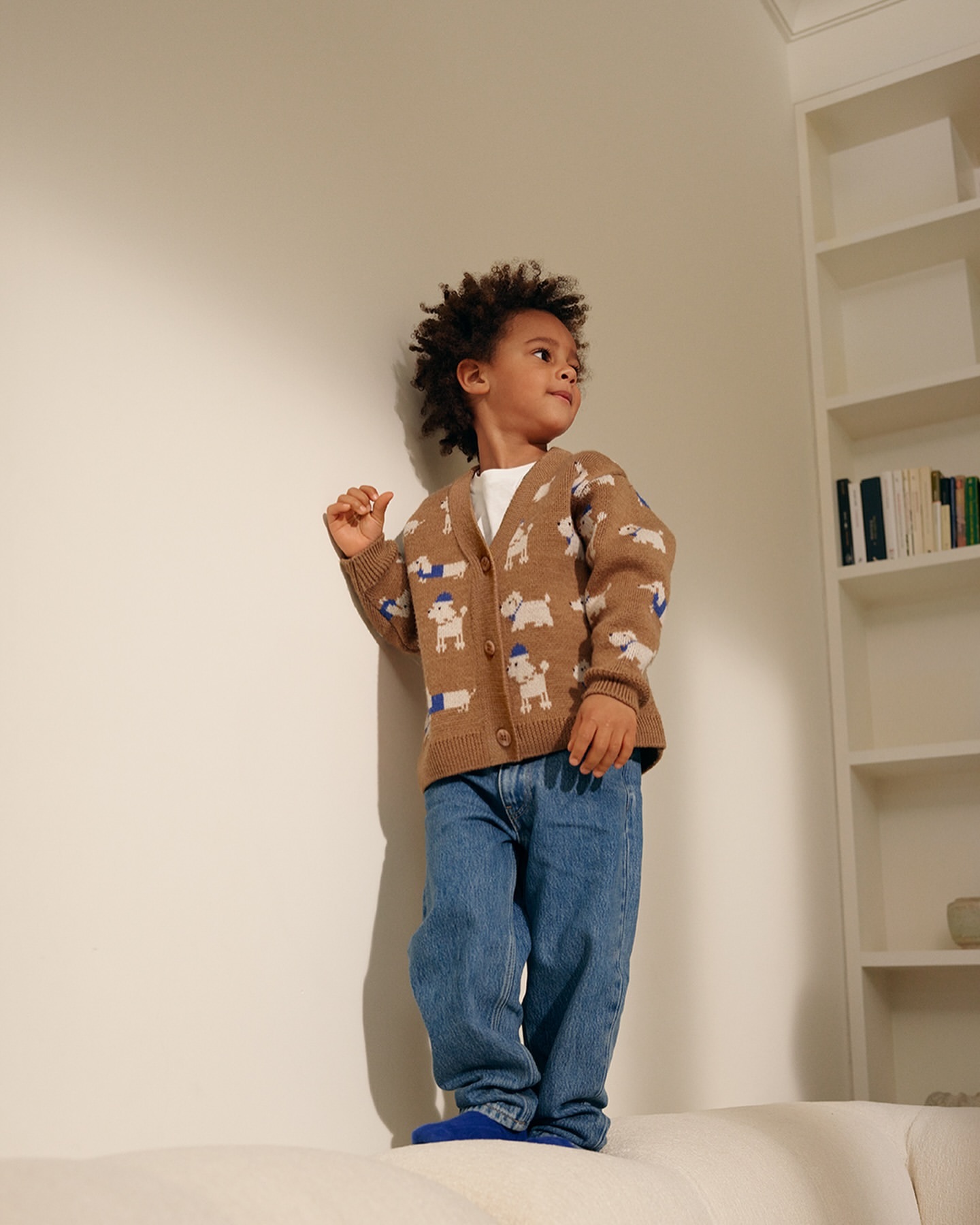 Young boy with curly hair stands on a white sofa in a tan cardigan with dog patterns, blue jeans, and blue socks in a bright indoor setting.