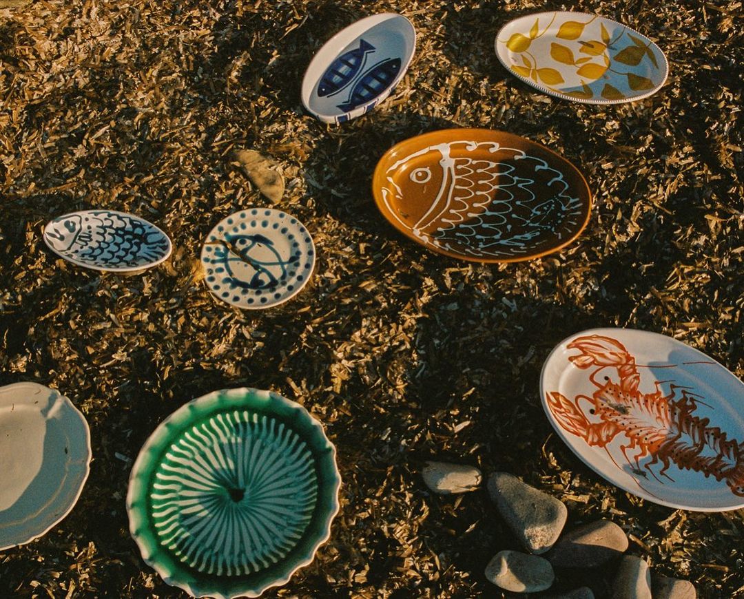 Overhead shot of variety of decorative ceramic plates with animal and leaf motifs, arranged on dry grass with some scattered pebbles.