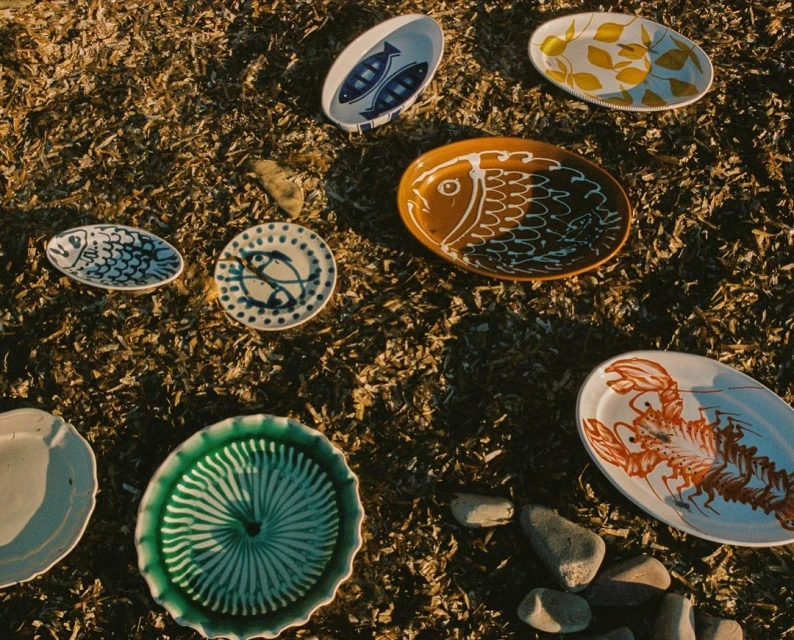 Overhead shot of variety of decorative ceramic plates with animal and leaf motifs, arranged on dry grass with some scattered pebbles.