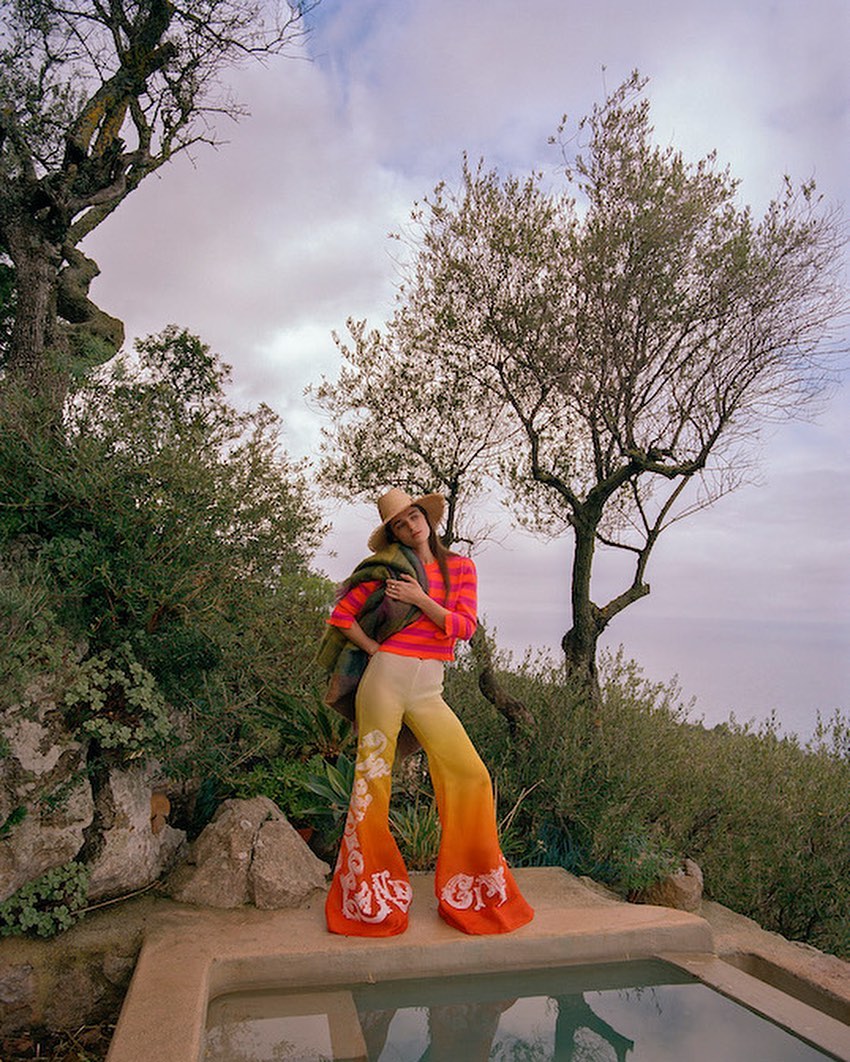 A fashionable woman in bell-bottom pants poses by a stone basin outdoors near a tree. She wears a hat and carries a jacket.