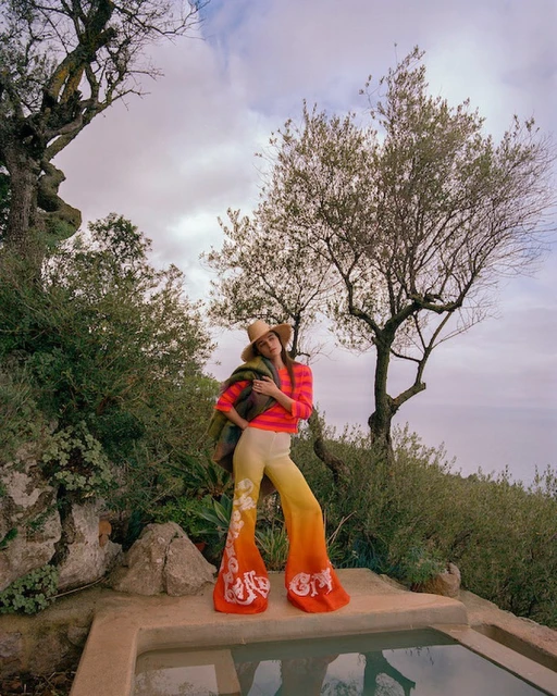 A fashionable woman in bell-bottom pants poses by a stone basin outdoors near a tree. She wears a hat and carries a jacket.