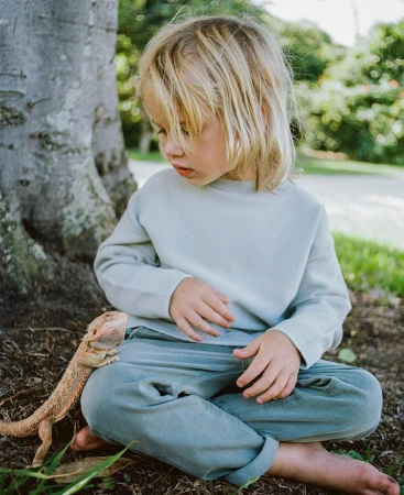 A young child with long blond hair sits barefoot on the grass outside near a tree with a pet bearded dragon lizard looking to the left.