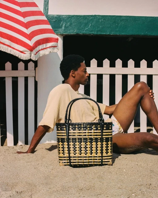 A Black man sitting on a sandy beach next to a woven beach bag with a sun umbrella and a beach house in the background.