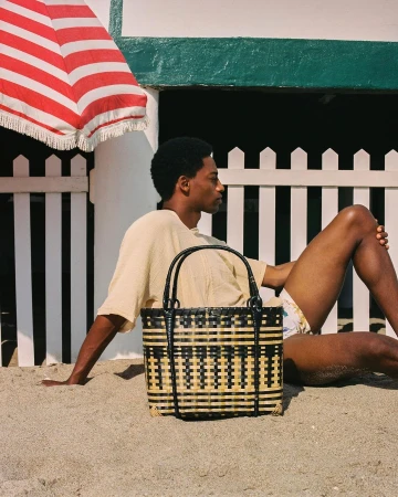 A Black man sitting on a sandy beach next to a woven beach bag with a sun umbrella and a beach house in the background.