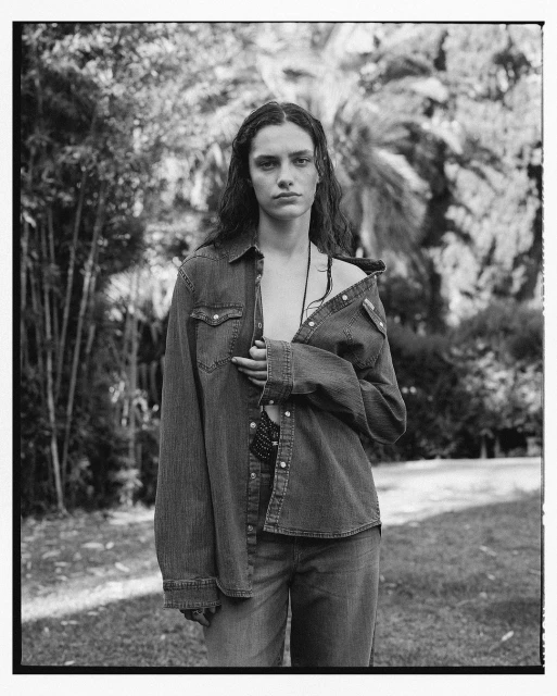 Monochrome portrait of a young woman with long hair, wearing a denim shirt and jeans, standing in a grassy outdoor environment.