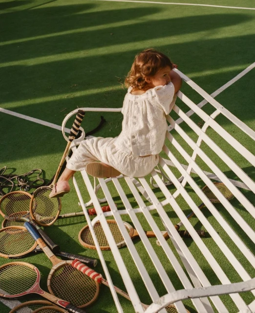 Young girl in cream-colored outfit rests on white bench amid tennis rackets on a green tennis court on a sunny day.