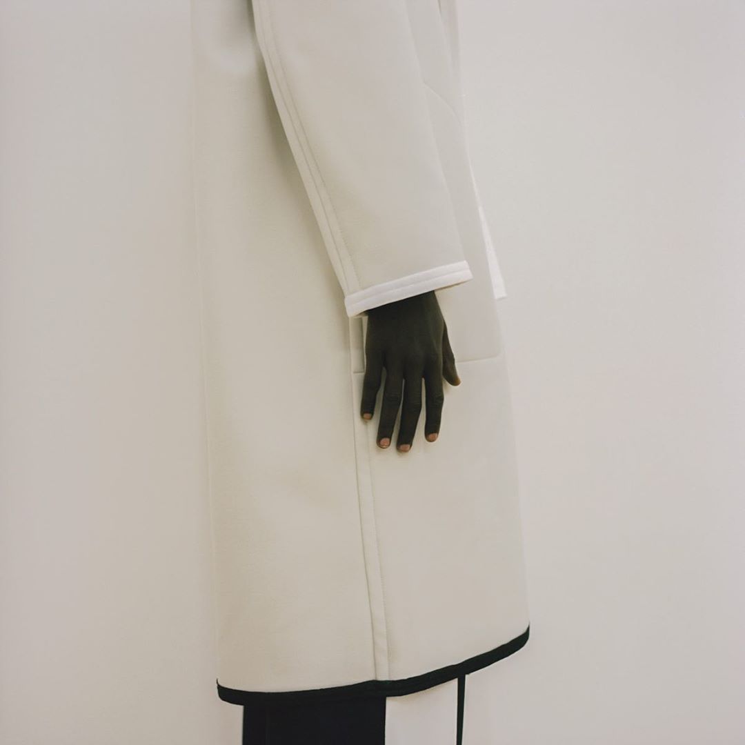 Fashion shot features a light coat with black trim, a dark hand, and black and white trousers against a bright, neutral background.