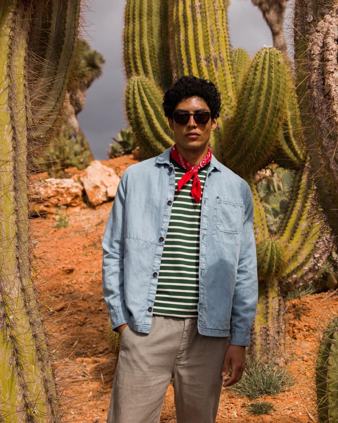 Man posing in front of cacti in a desert setting, wearing a denim shirt, striped shirt, khaki pants, sunglasses, and a red bandana.