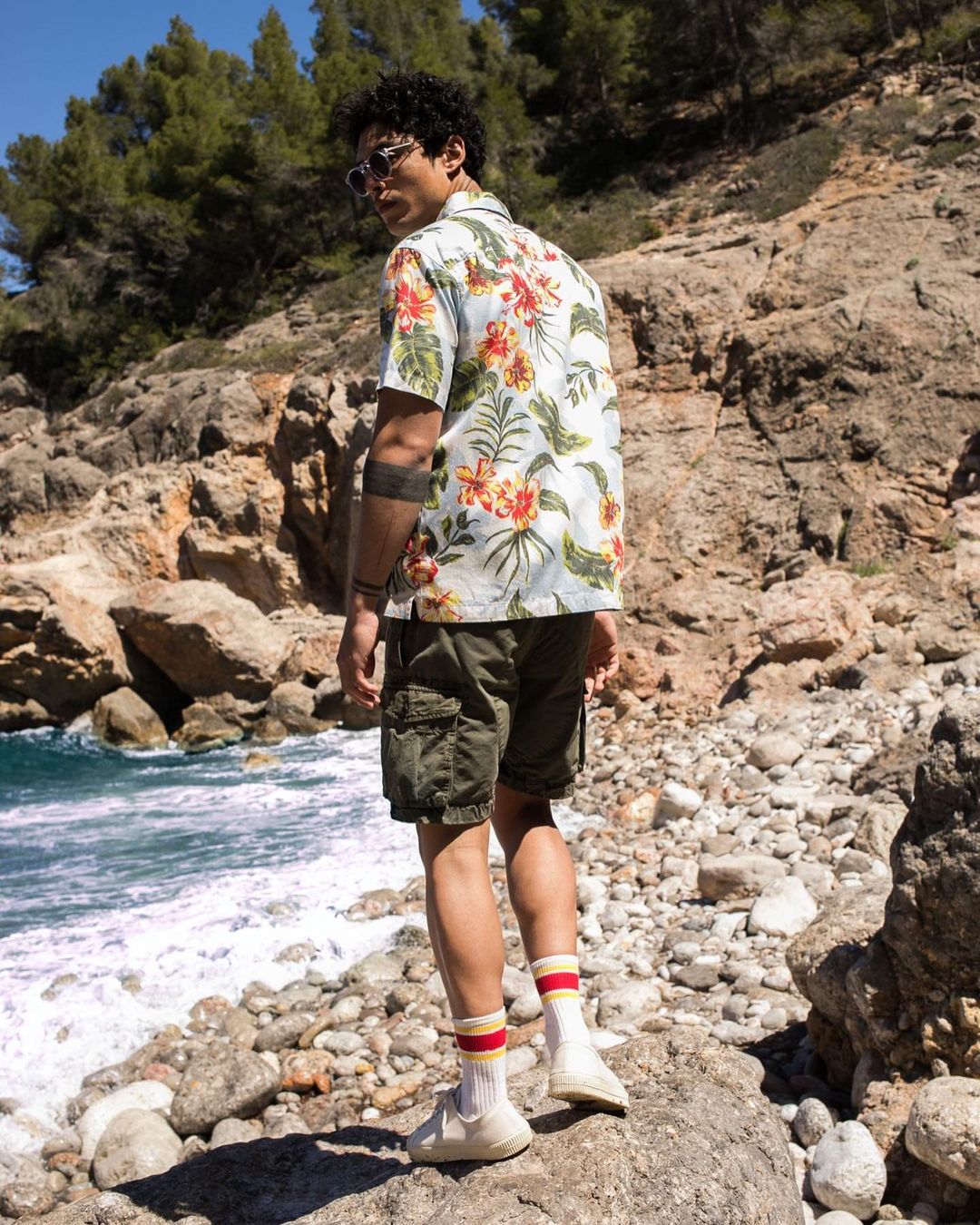 Man wearing floral shirt, olive shorts, sunglasses poses on rocky beach with ocean and rock formations in outdoor daytime shot.
