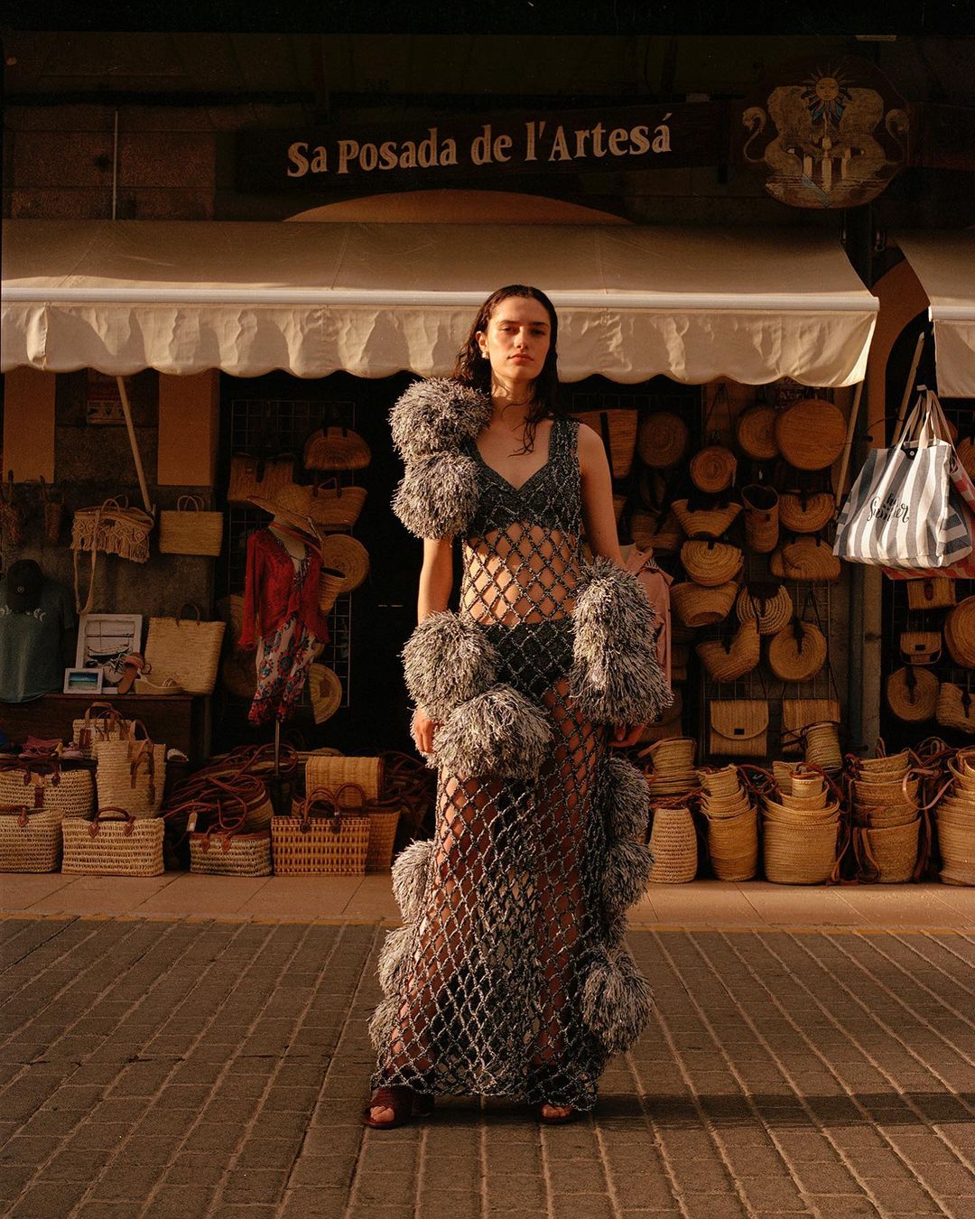 A woman in a fashion dress poses outdoors in front of a store with straw baskets, under warm lighting on a brick path.