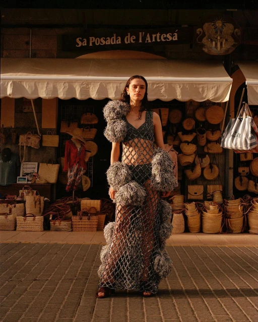 A woman in a fashion dress poses outdoors in front of a store with straw baskets, under warm lighting on a brick path.