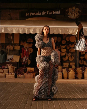 A woman in a fashion dress poses outdoors in front of a store with straw baskets, under warm lighting on a brick path.