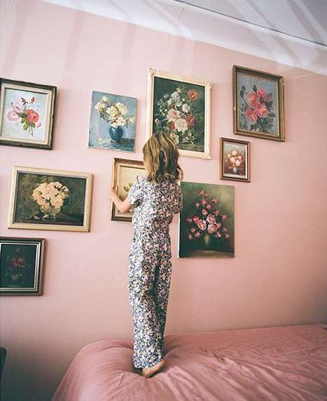 Young child in patterned pajamas stands on a bed, hanging floral painting on a pink wall adorned with various framed flower artworks.