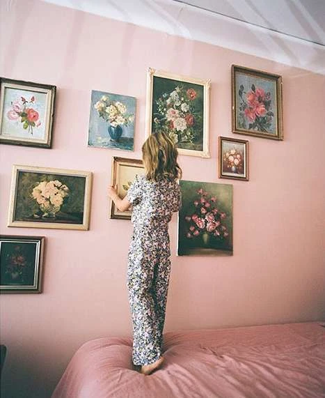 Young child in patterned pajamas stands on a bed, hanging floral painting on a pink wall adorned with various framed flower artworks.