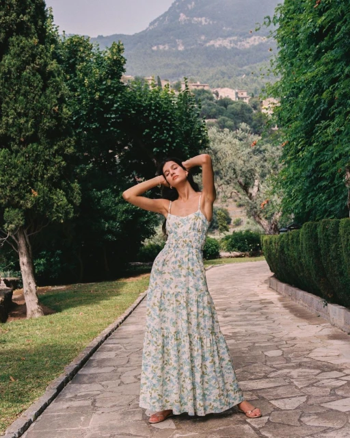 Woman in floral maxi dress standing on a stone path surrounded by greenery, with mountains in the background, creating a scenic outdoor fashion shot.