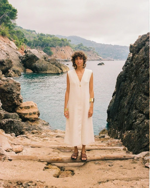 Woman in a long dress standing on a rocky path near the ocean, with a mountain backdrop, fashion shoot, natural light, brown sandals.
