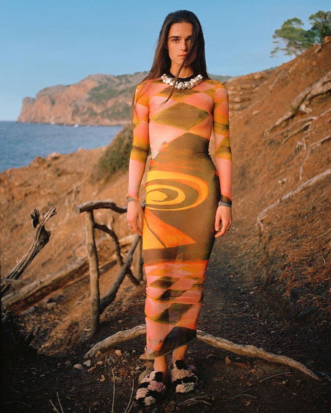 Adult woman wearing a patterned dress poses on a dirt path near a rocky coastline on a sunny day in an eye-level medium shot.