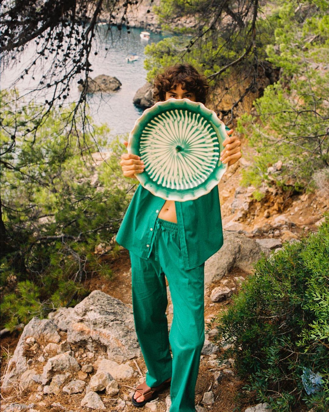 Young woman in a green outfit holds a round, patterned plate in front of her face on a rocky path near the Mediterranean coast.