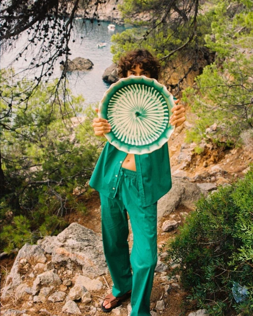 Young woman in a green outfit holds a round, patterned plate in front of her face on a rocky path near the Mediterranean coast.