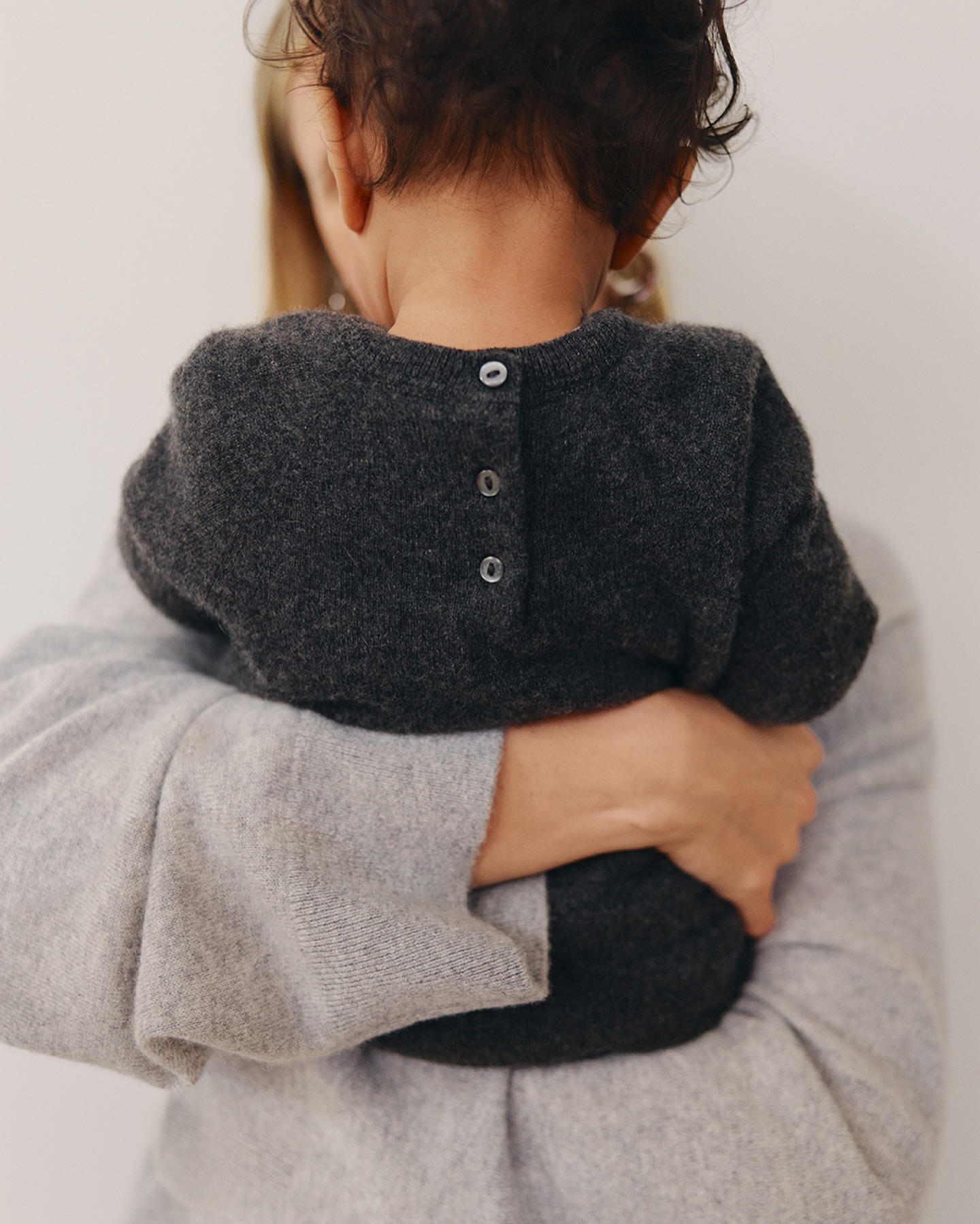 A child in a grey sweater is held by a woman, both facing away from the camera, with a bright, neutral background, and soft lighting.