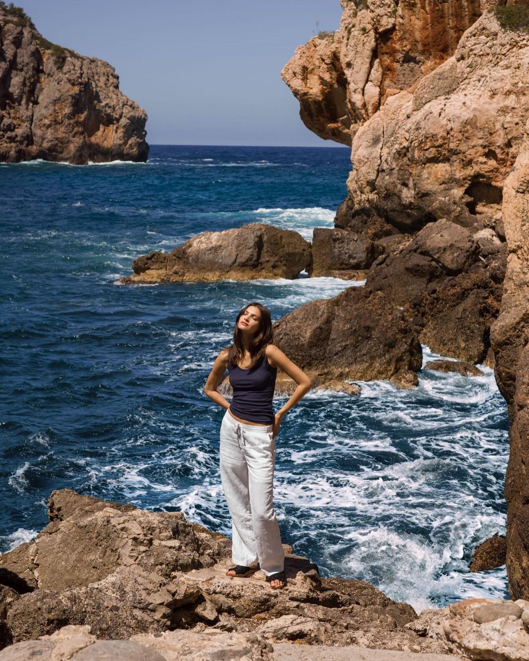 Young woman stands on rocks overlooking ocean with rocky cliffs and clear blue sky in outdoor, natural light fashion shot.