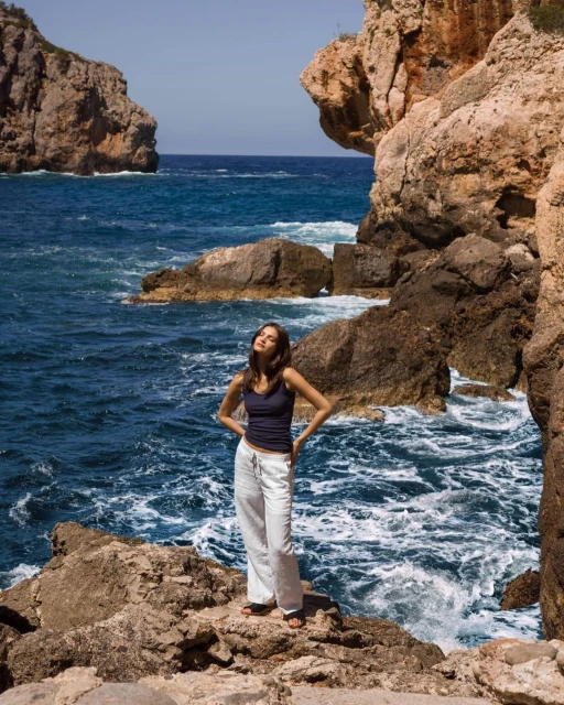 Young woman stands on rocks overlooking ocean with rocky cliffs and clear blue sky in outdoor, natural light fashion shot.