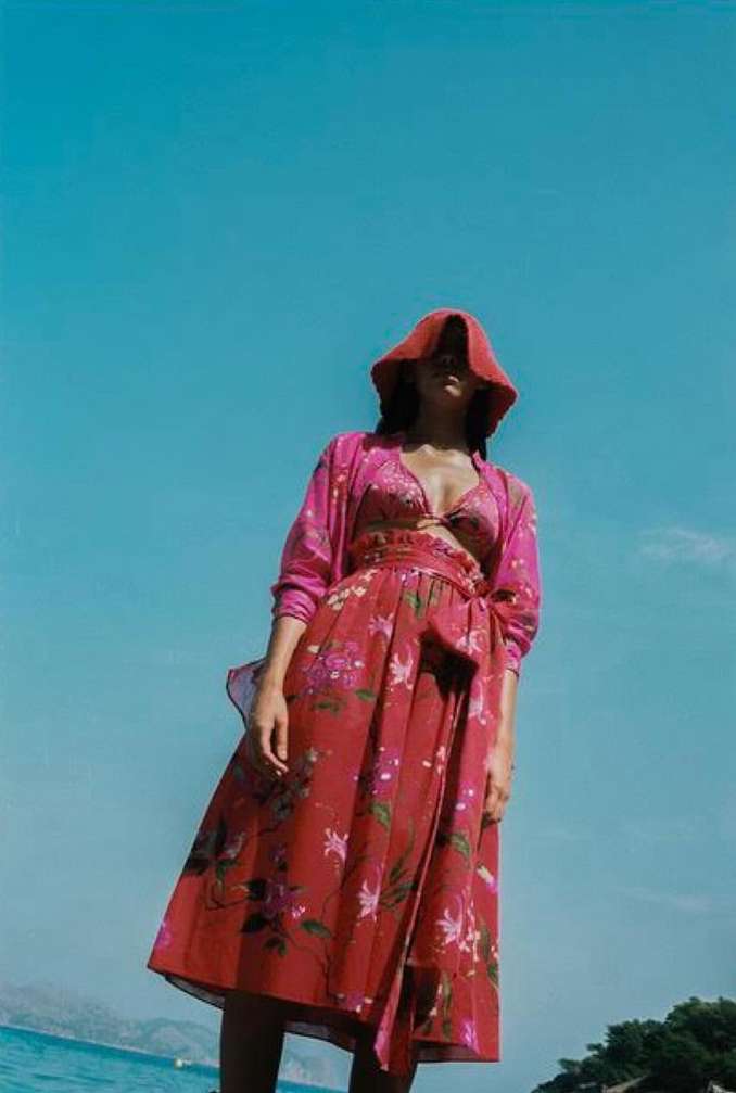 A woman in a pink floral dress and hat stands against a blue sky and ocean backdrop, showcasing a beach fashion editorial.