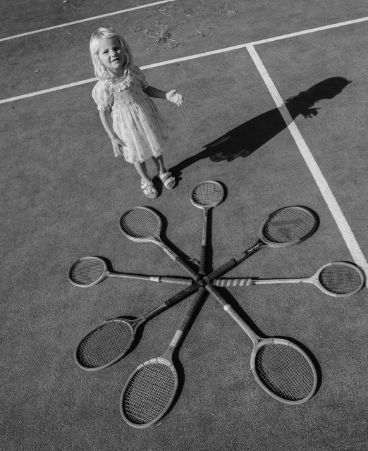 Young girl stands next to a pinwheel design of tennis rackets on an outdoor tennis court, captured in a black and white, overhead shot.