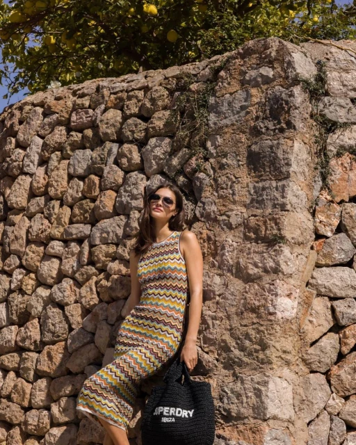 A woman in a zig-zag patterned dress and sunglasses stands near a stone wall, holding a black tote bag. Lemon tree in the background.