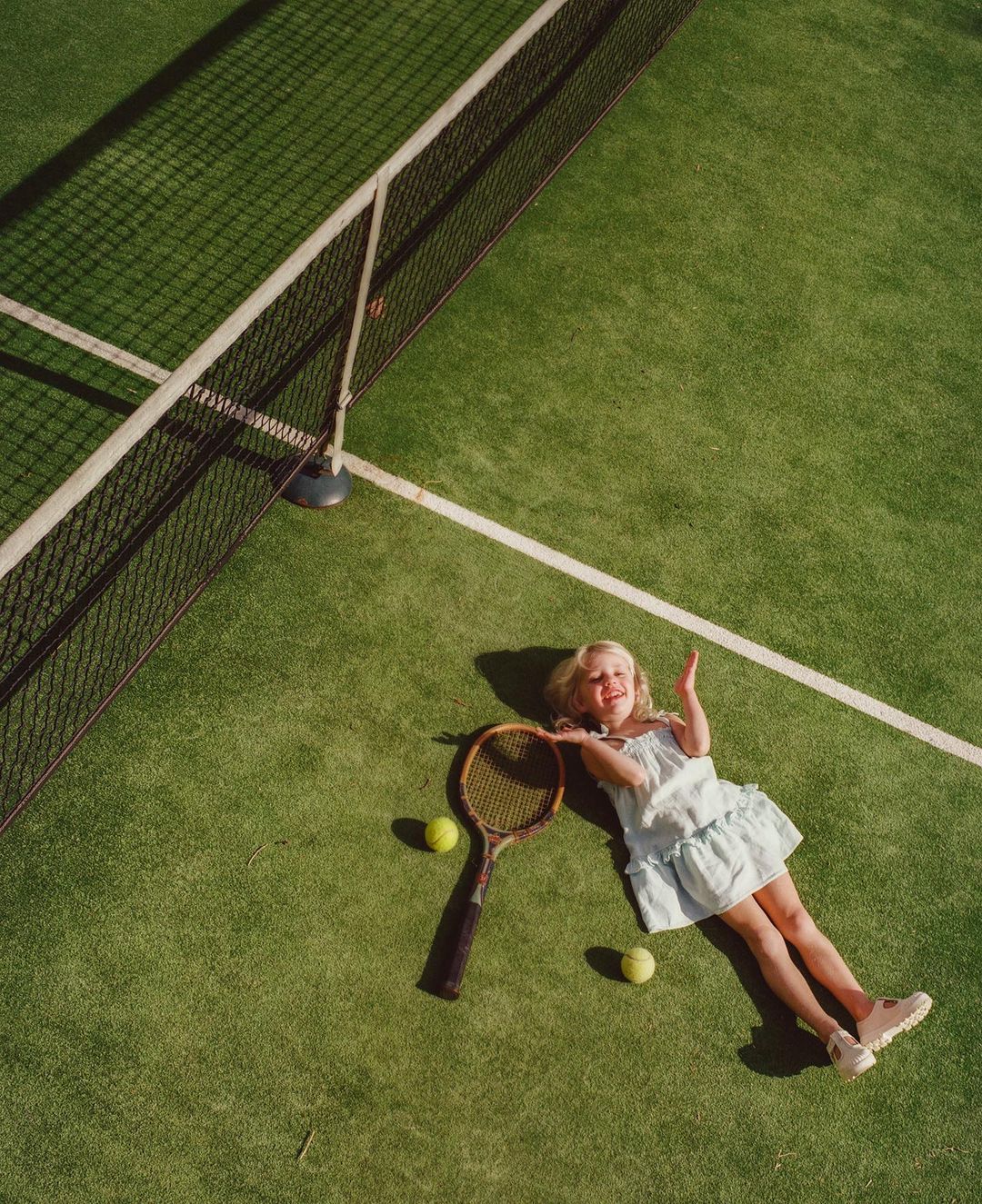 Overhead view of a blonde child lying on a green tennis court with a racket, tennis balls, and white lines, creating a playful and sporty scene.