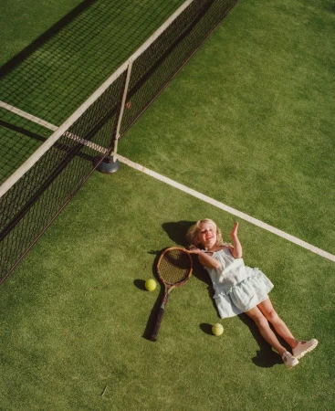 Overhead view of a blonde child lying on a green tennis court with a racket, tennis balls, and white lines, creating a playful and sporty scene.